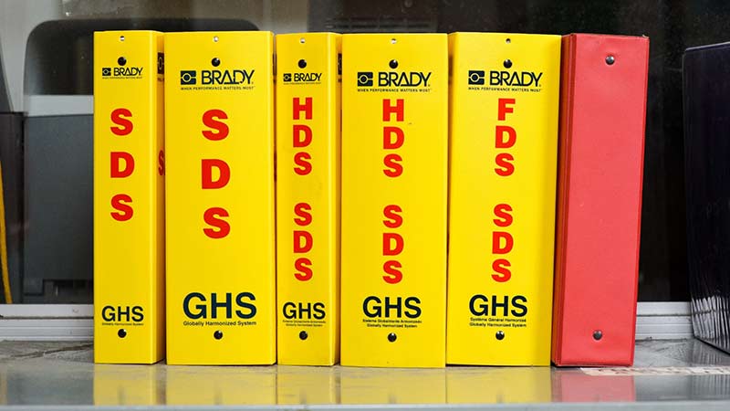 Four yellow SDS binders sitting on a table below a poster detailing OSHA's HazCom information.